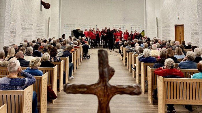 Et moderne kirkeinteriør med siddende menighed, kor der optræder, og en fremtrædende træ kors i forgrunden.