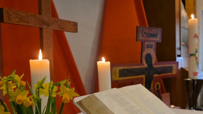 Ruhiger Altar mit offenem Buch, leuchtenden Blumen und brennenden Kerzen unter einem hölzernen Kreuz.