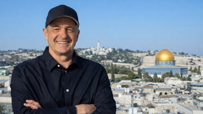Ein lächelnder Mann mit einer Mütze steht selbstbewusst mit verschränkten Armen und blickt auf Jerusalems Felsendom.