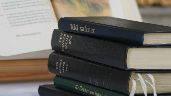 En stabel af bøger på seng, med en åben bog i baggrunden