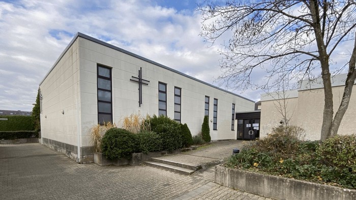 Moderne Kirchengebäude mit einem Kreuzsymbol an der Fassade, umgeben von gestalteter Grünanlage.