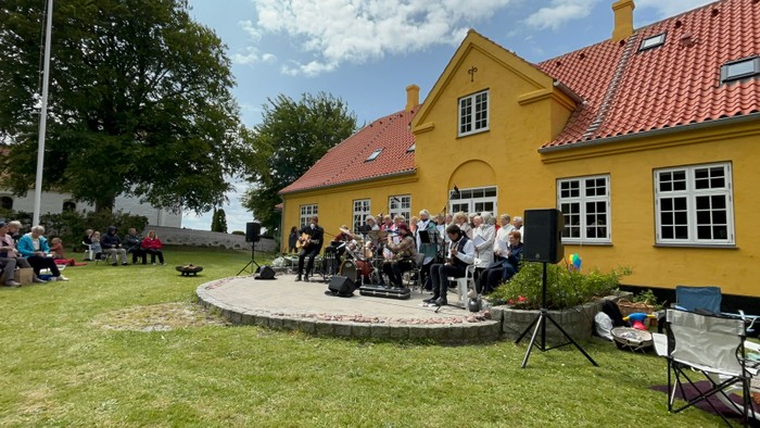 Udendørs musikoptræden med et orkester på en græsklædt scene nær en gul bygning.