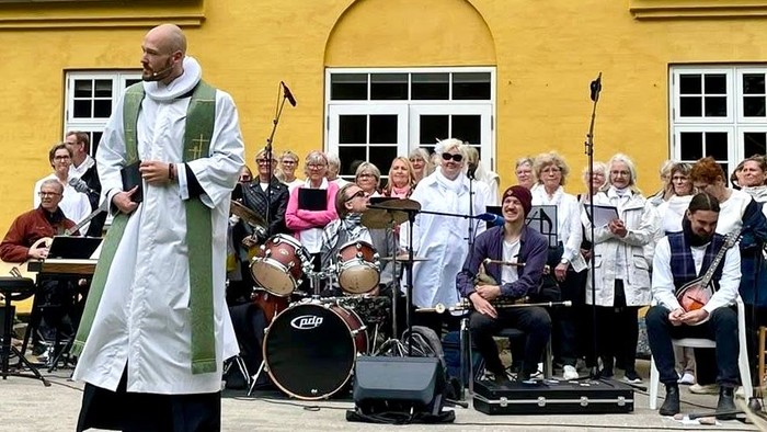 En præst står og taler til en folkemængde foran et gult hus med en musikgruppe bagved.