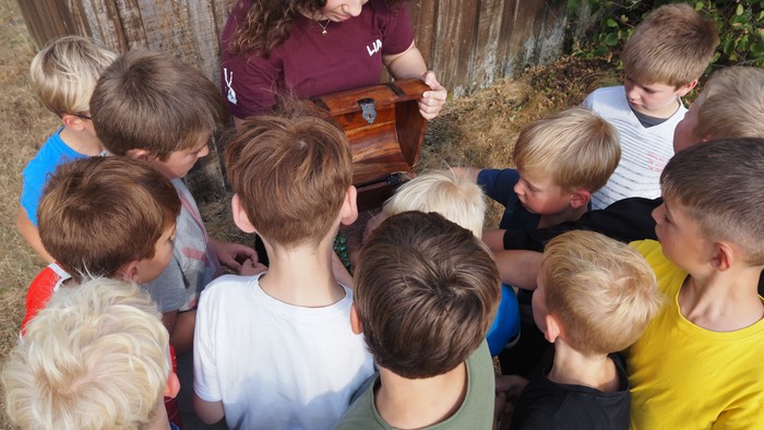Eine Gruppe von Kindern versammelt sich aufmerksam um einen Erwachsenen, der draußen etwas an einer kleinen Holzkiste vorführt.