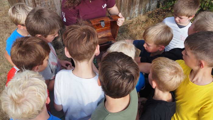Eine Gruppe von Kindern versammelt sich aufmerksam um einen Erwachsenen, der draußen etwas an einer kleinen Holzkiste vorführt.