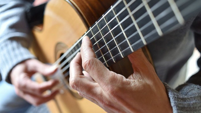 Die Nahaufnahme zeigt die Hände einer Person beim Spielen einer akustischen Gitarre. Im Vordergrund ist die linke Hand zu sehen, die die Saiten auf dem Griffbrett greift und einen Akkord formt. Die Finger liegen präzise zwischen den Bundstäbchen, während 