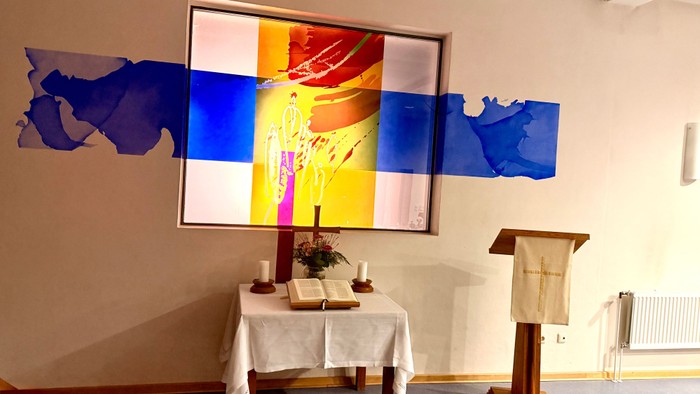 Religiöser Altarraum mit lebendigem Wandgemälde, Kerzen, Büchern und einem Lesepult in einer ruhigen Innenraumatmosphäre.