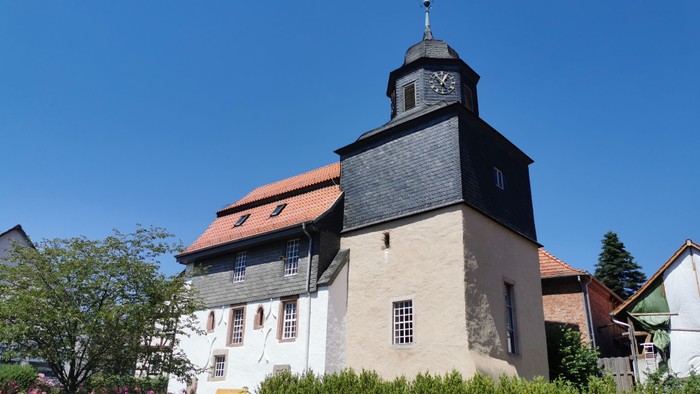Das Bild zeigt eine Kirche mit Uhrturm und roter Ziegeldach, umgeben von grünem Gestrüpp unter klarem Himmel.