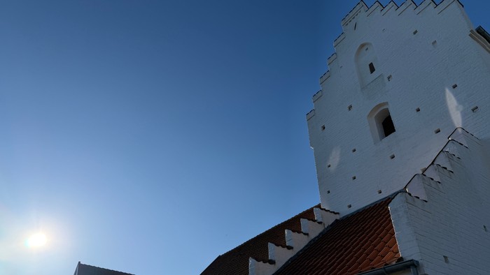 En hvid kirkebygning med rødt tag på en solskinsdag med blå himmel.