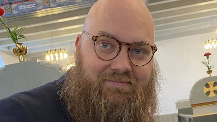 En skægget mand med briller står indendørs nær religiøse ikoner og glasmosaikvinduer.