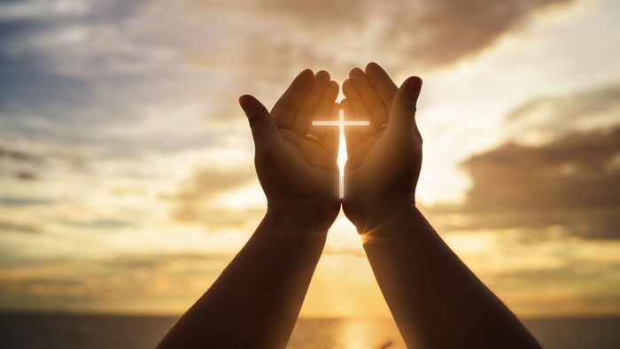 Betende Hände mit Kreuz im Gegenlicht eines Sonnenaufgangs