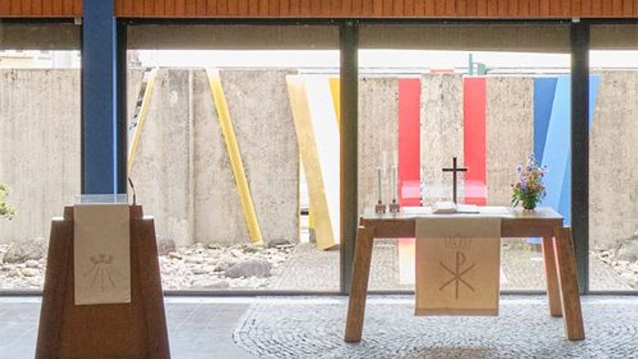 Minimalistische Outdoor-Kapellen-Einrichtung mit hölzernen Podesten, religiösen Symbolen und farbenfrohen geometrischen Wänden im Hintergrund.