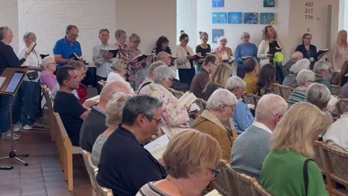 En gruppe mennesker samlet i et rum, nogle siddende og nogle stående, engageret i en musikalsk aktivitet med nodeark og instrumenter.