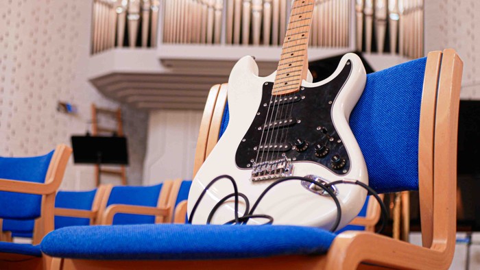 En hvid elektrisk guitar hviler på en blå polstret stol i et rum med træbænke og orgelpiber.