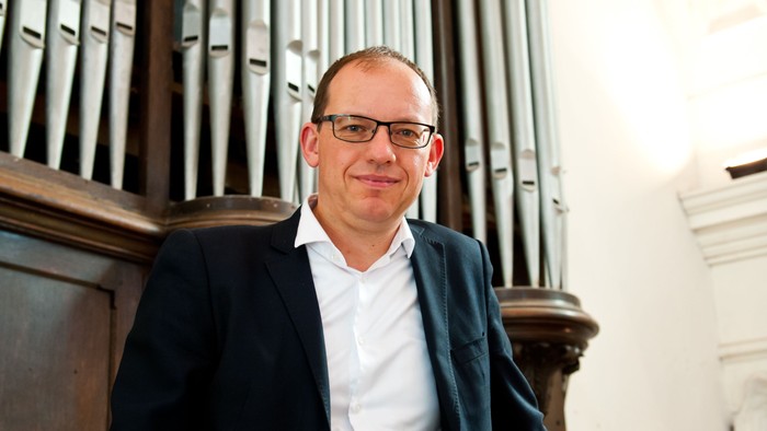 En mand med briller og en blazer læner sig op ad et stort orgel i en kirke.