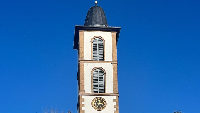 Kirchturm mit Uhr und Kreuz auf dem Dach, umgeben von Bäumen und Himmel