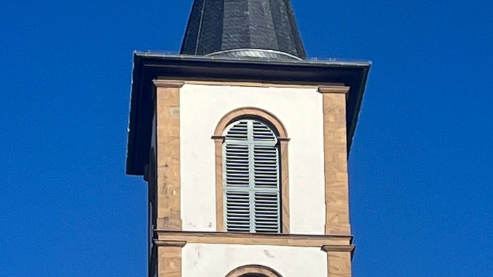 Kirchturm mit Uhr und Kreuz auf dem Dach, umgeben von Bäumen und Himmel
