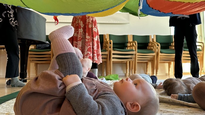 Et spilende barn ligger på ryggen under en farverig paraply i legeværelset