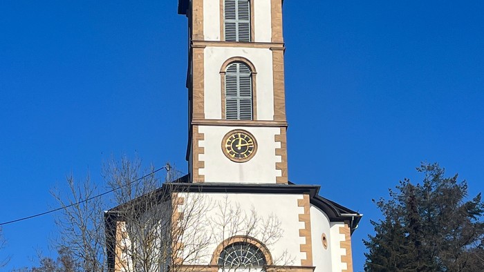 Kirchturm mit Uhr und Kreuz auf dem Dach, umgeben von Bäumen und Himmel