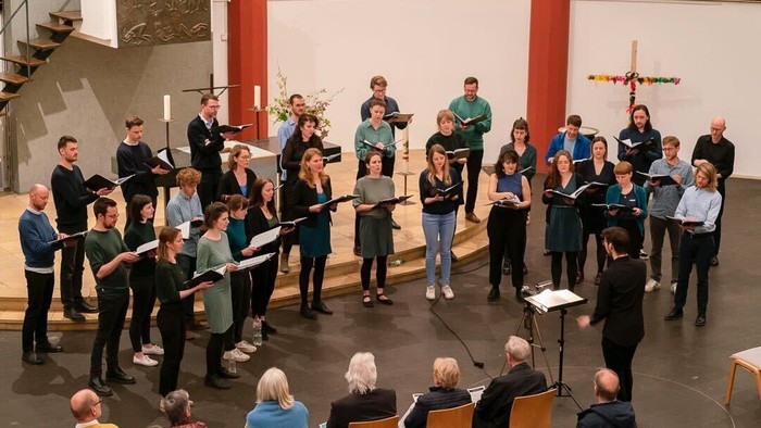 Ein Chor probt in einem großen Raum mit hohen Decken und einer Treppe im Hintergrund.