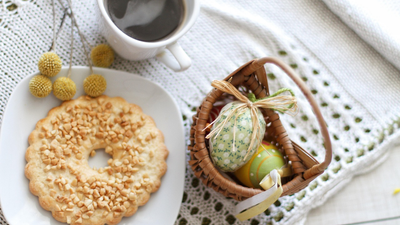 Eine gemütliche Anordnung mit einem Donut, Kaffee, dekorativen Eiern und einem Korb auf einer Spitzen-Tischdecke.