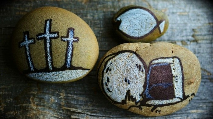 Udsmykkede glatte sten, der symboliserer kors, fisk og et tomt gravkammer på en træoverflade.