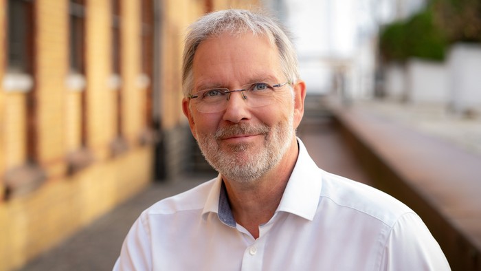Ein lächelnder Mann mit Brille und weißem Hemd, der draußen in der Nähe eines Gebäudes steht.