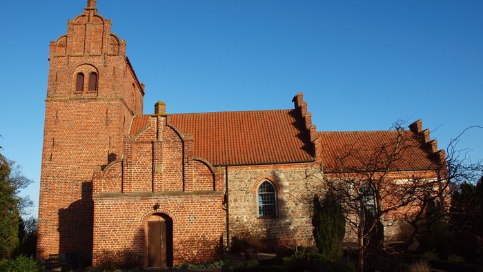 En gammel teglmuret kirke med klokketårn og rødt tag, omgivet af træer og buske
