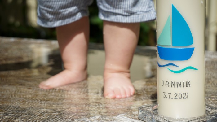 Füßchen eines Babys neben einer Kerze mit Segelboot-Motiv und dem Namen „Jannik 3.7.2021“.