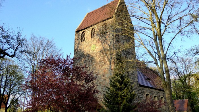 Alte Kirche mit hohem Turm und rotem Dach, umgeben von Bäumen und Büschen