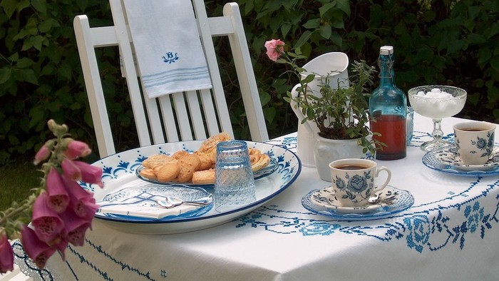 Idyllisches Outdoor-Teetisch-Arrangement mit Gebäck, Blumen und altem Porzellan auf einer blauen Tischdecke.