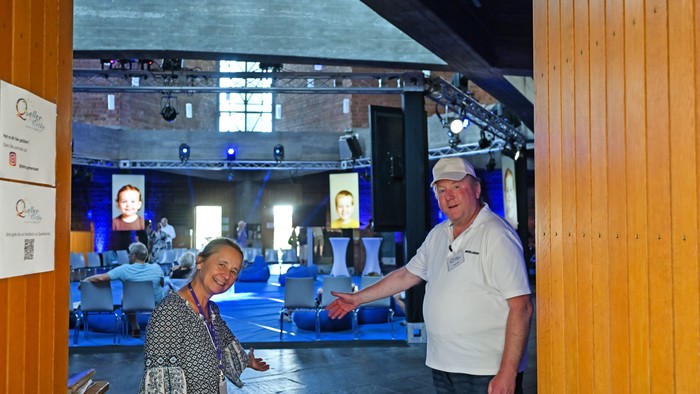 Zwei Personen geben sich in einem geräumigen Veranstaltungsort mit Bühne und Publikumsbestuhlung im Hintergrund die Hand.