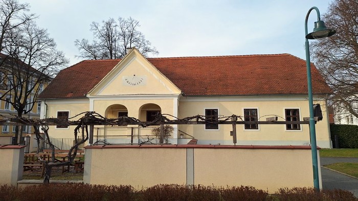 Das Bild zeigt ein Gebäude mit rotem Dach und weißen Wänden, umgeben von einem Zaun und Bäumen.