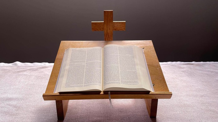 Geöffnete Bibel auf einem hölzernen Lesepult mit einem Kreuz darauf, platziert auf einem weißen Tuch.