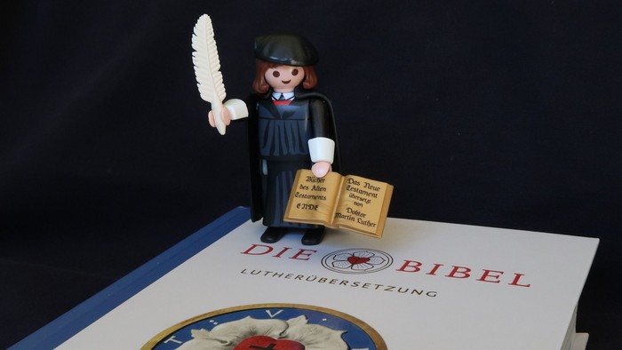 Eine Playmobil-Figur hält eine Feder und ein Buch, auf einem Buch mit einem blauen Einband und einem roten Käfer.