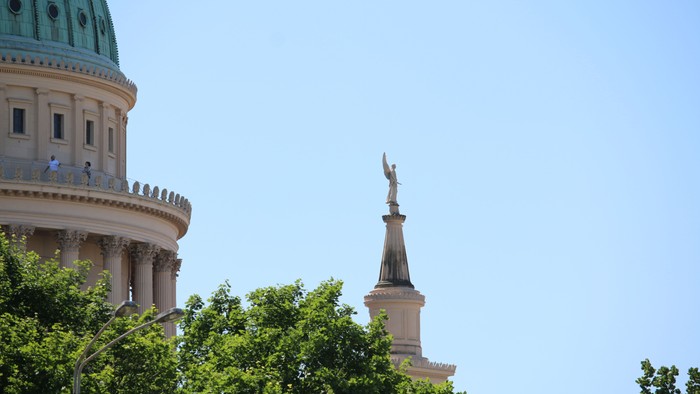 Das Bild zeigt eine Kuppel mit grüner Kuppel und eine Statue darauf, umgeben von Bäumen