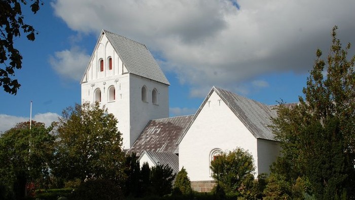 En hvid kirke med tårn og spids tak, omgivet af træer og græs.