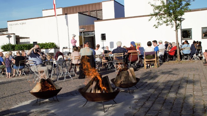 En gruppe af mennesker sidder udendørs ved firebåle foran et hvidt hus.