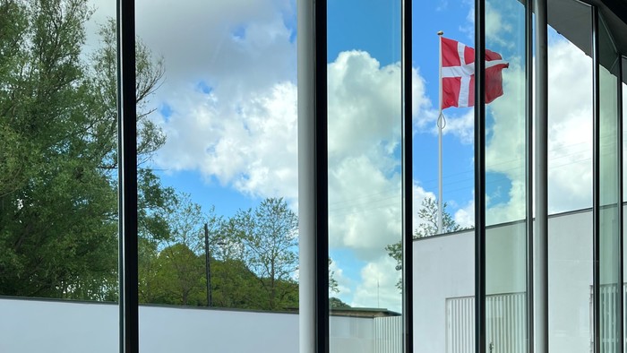 En stor vinduesfront med udsigt over vand og træer, med en plante i forgrunden og et flag i baggrunden