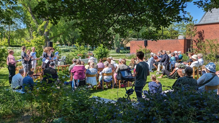 En gruppe af mennesker sidder og står i en have med en rød bygning i baggrunden