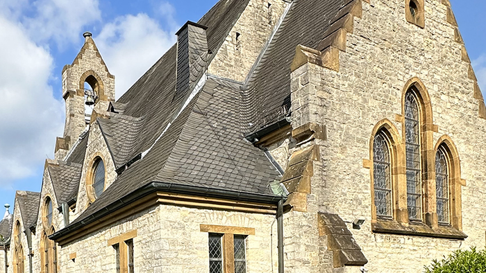 Historische Steinkirche mit Spitzbögen, steilem Dach und angrenzenden Bäumen unter einem teilweise bewölkten Himmel.