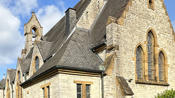 Historische Steinkirche mit Spitzbögen, steilem Dach und angrenzenden Bäumen unter einem teilweise bewölkten Himmel.