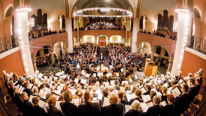 Ein großer Konzertsaal, gefüllt mit einem Publikum und einem Orchester, das auf der Bühne spielt.