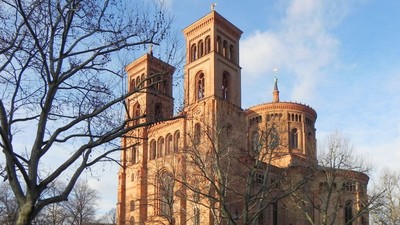 Großes, altes Backsteingebäude mit zwei Türmen und Rundbau, umgeben von kahlen Bäumen und einem Auto