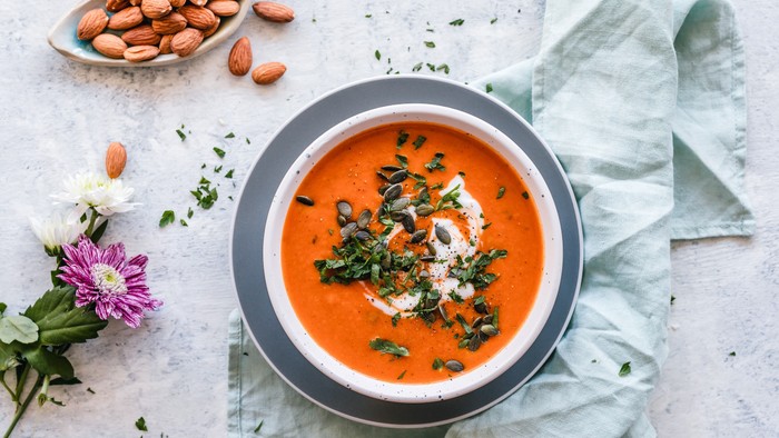 Warme, cremige Tomatensuppe, garniert mit Grünzeug, Mandeln und frischen Kräutern neben einer Schüssel.