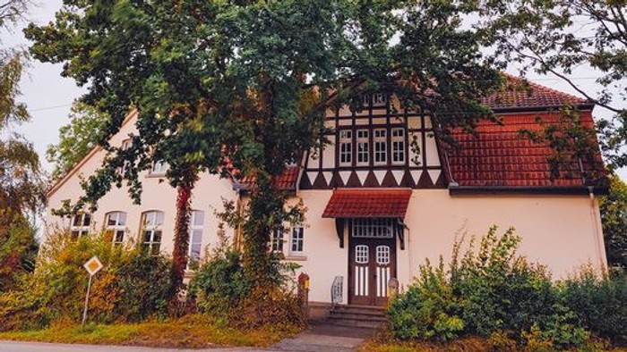 Das Bild zeigt ein charmantes, traditionelles Haus mit einem roten Dach und weißen Wänden, umgeben von üppigen grünen Bäumen und Büschen.