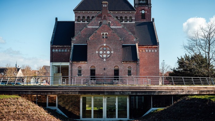 Et rødt teglsteinshus med spids tak og en stor hjerteformet vindueskarm på fasaden. Foran huset er der en bro over en grøft.