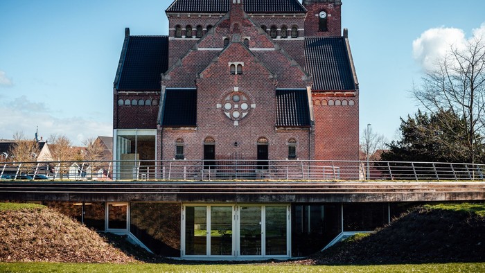 Et rødt teglsteinshus med spids tak og en stor hjerteformet vindueskarm på fasaden. Foran huset er der en bro over en grøft.