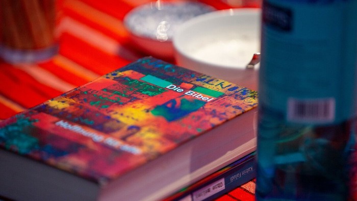 Im Vordergrund liegt eine Bibel mit farbenfrohem Einband auf einem Tisch mit rot gestreifter Tischdecke. Auf dem Cover ist deutlich der Titel „Die Bibel“ zu lesen. Das Buch liegt auf weiteren Büchern oder Heften. Im Hintergrund stehen unscharf eine Schal
