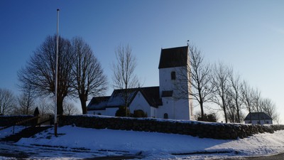 En hvid kirke med tårn og et mindre sidebygning, omgivet af sne og nakke træer.